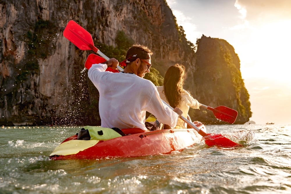 young couple paddles through phang nga bay in a red kayak with limestone cliffs nearby